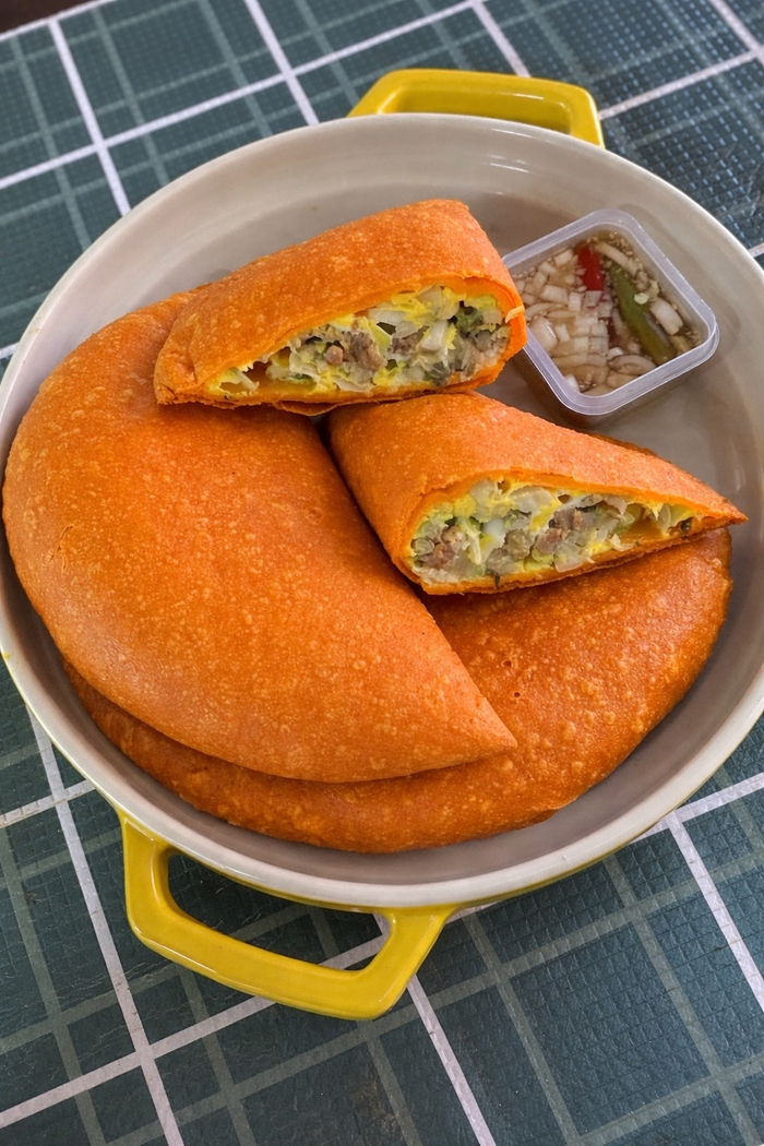 Ilocos empanadas with a bite taken out of one and a side of spiced vinegar, served in a white bowl with yellow handles on a checkered tablecloth.
